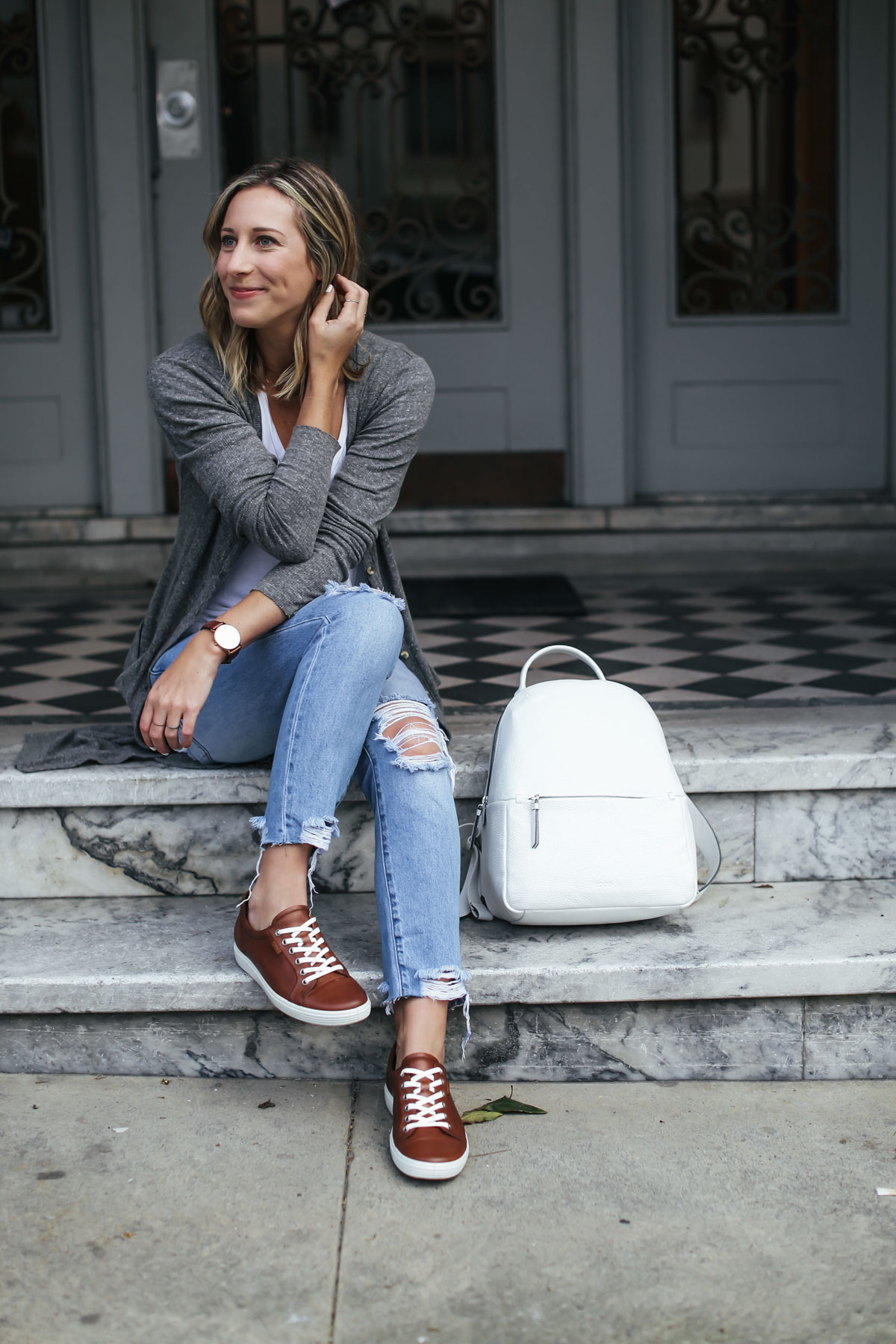 travel outfit with distressed boyfriend jeans, white leather backpack, brown leather sneakers, and gray cardigan