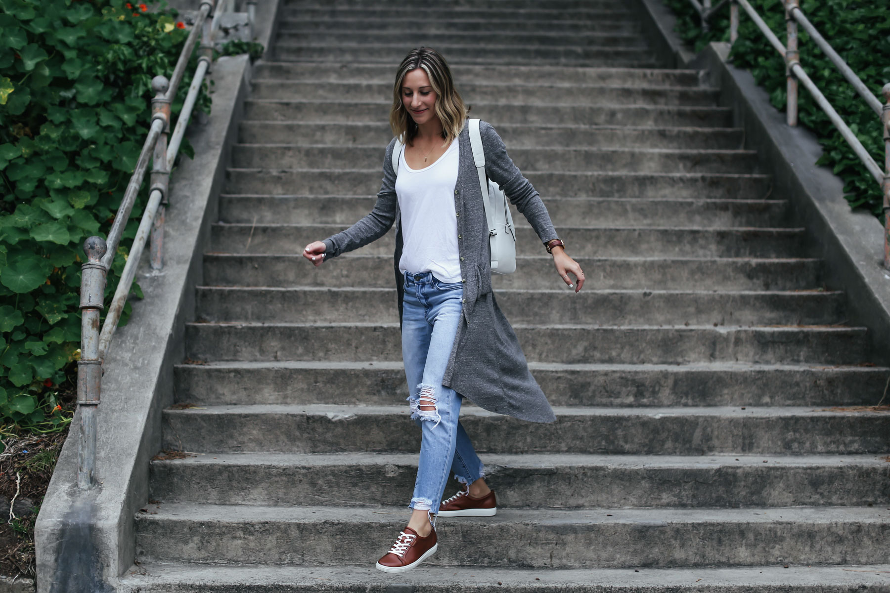 travel outfit with distressed boyfriend jeans, white leather backpack, brown leather sneakers, and gray cardigan